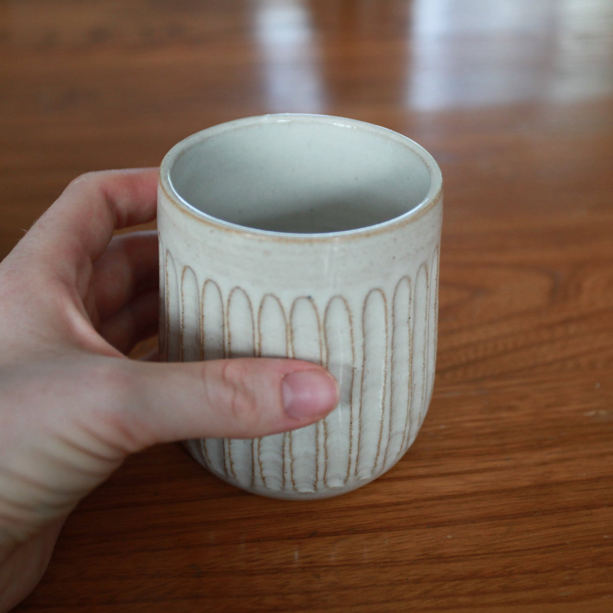 A hand holding a small ceramic mug with a ribbed, scalloped exterior and a light gray interior, set against a warm wooden tabletop background.