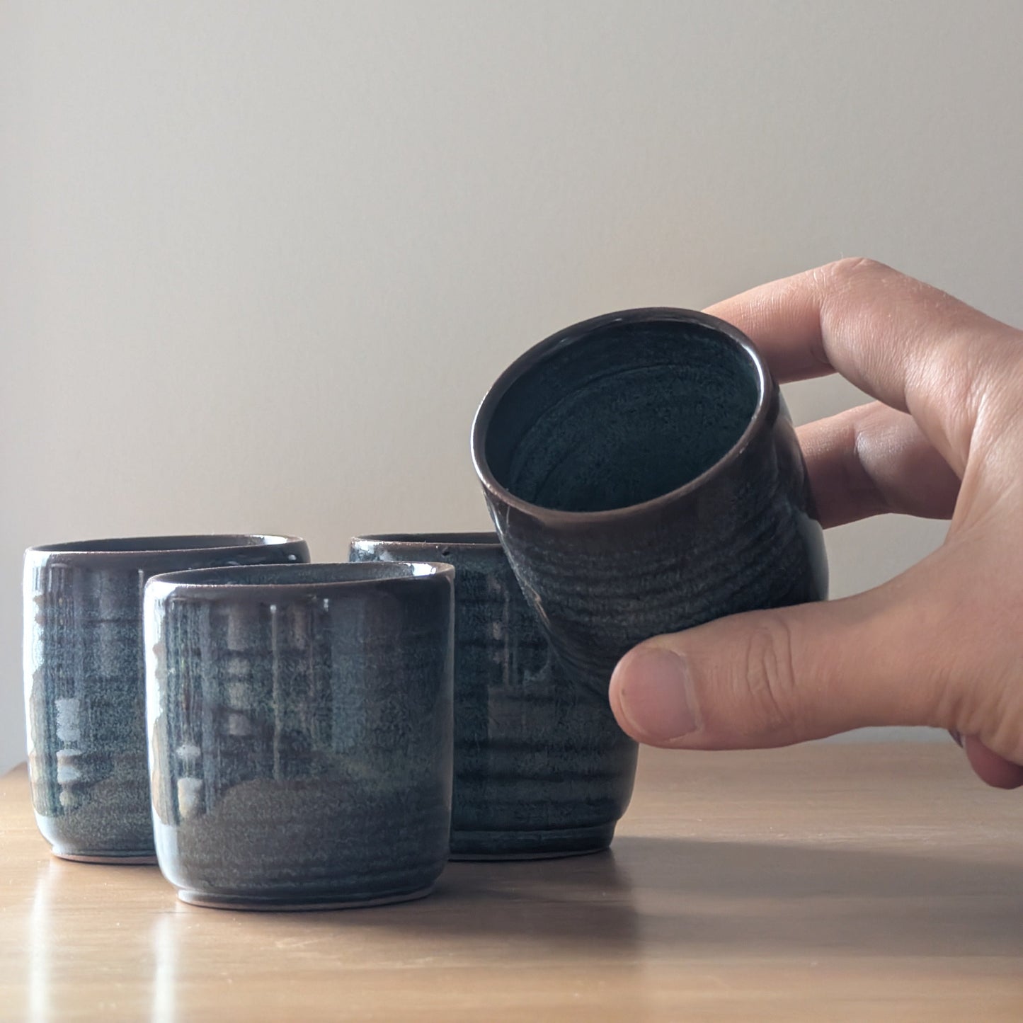 Hand holding a dark ceramic cup with other similar cups on a wooden surface.