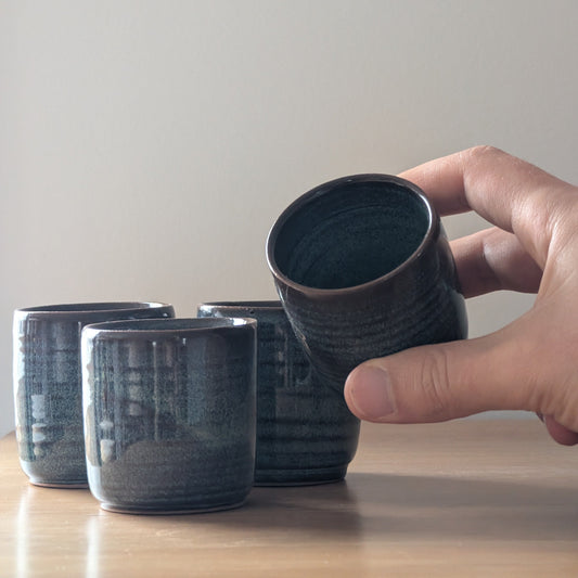 Hand holding a dark ceramic cup with other similar cups on a wooden surface.